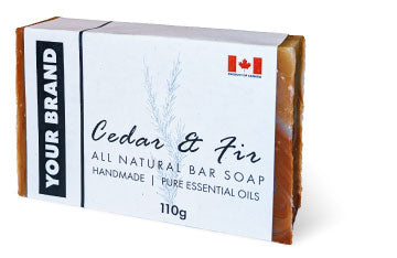A bar of Cedar & Fir all-natural handmade soap. Its white label features an illustration of a fir sprig, a small Canadian flag icon, and a side panel with a placeholder for 'YOUR BRAND'.